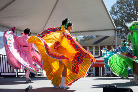 three Ballet Folklorico Dancers on stage, wearing pink, yellow and green dresses